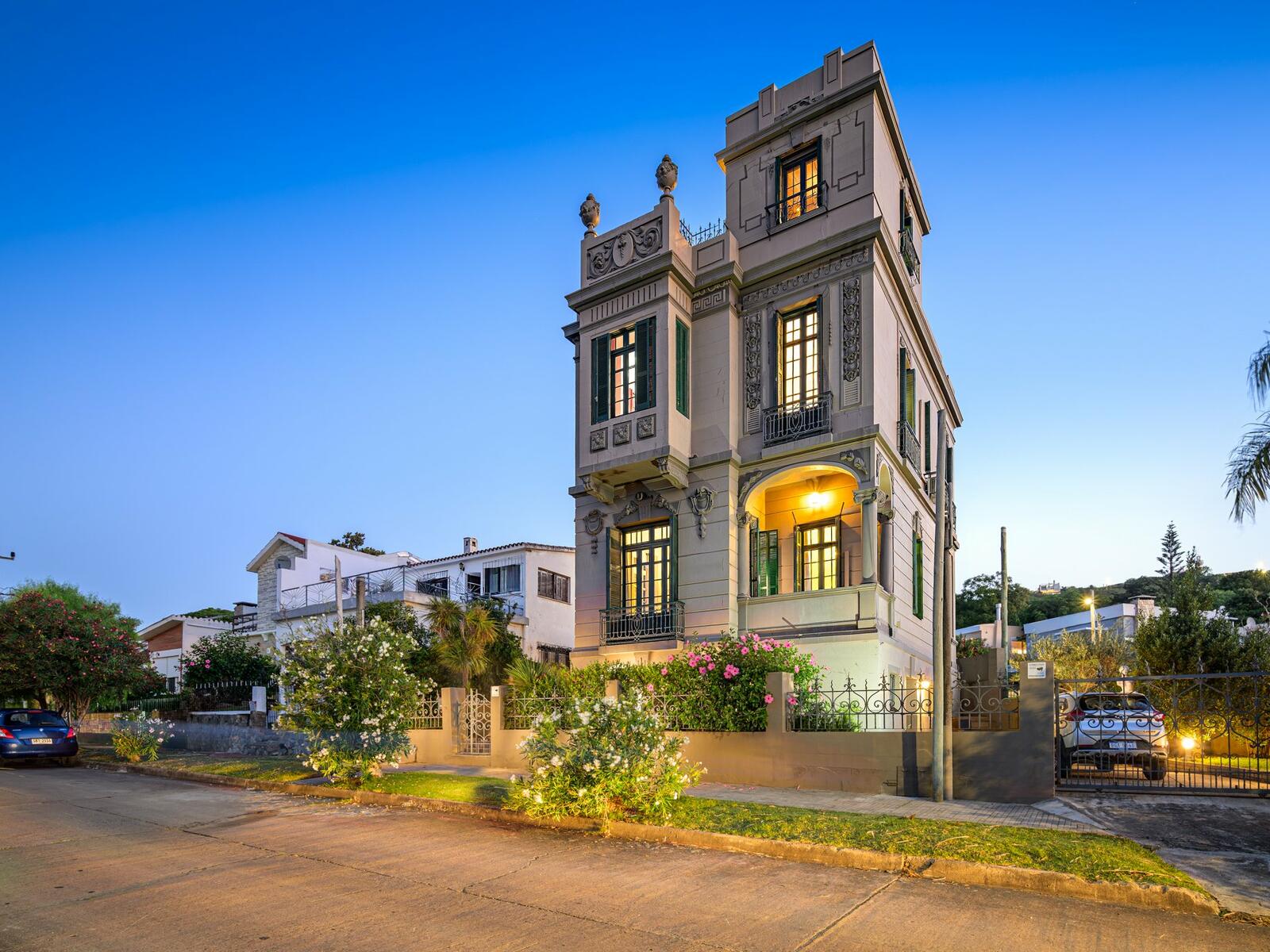Restored 1912 heritage home in Piriápolis photographed at dusk, with the tower and full facade lit up above the street